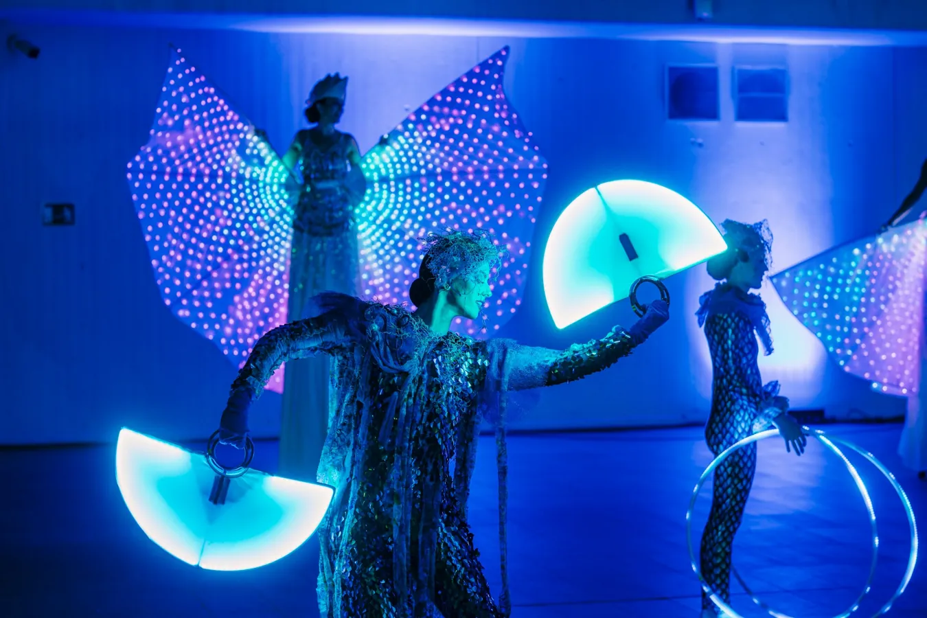 Tres artistas con grandes alas luminosas actúan en la Terraza de L’Aquàrium durante un evento al atardecer.