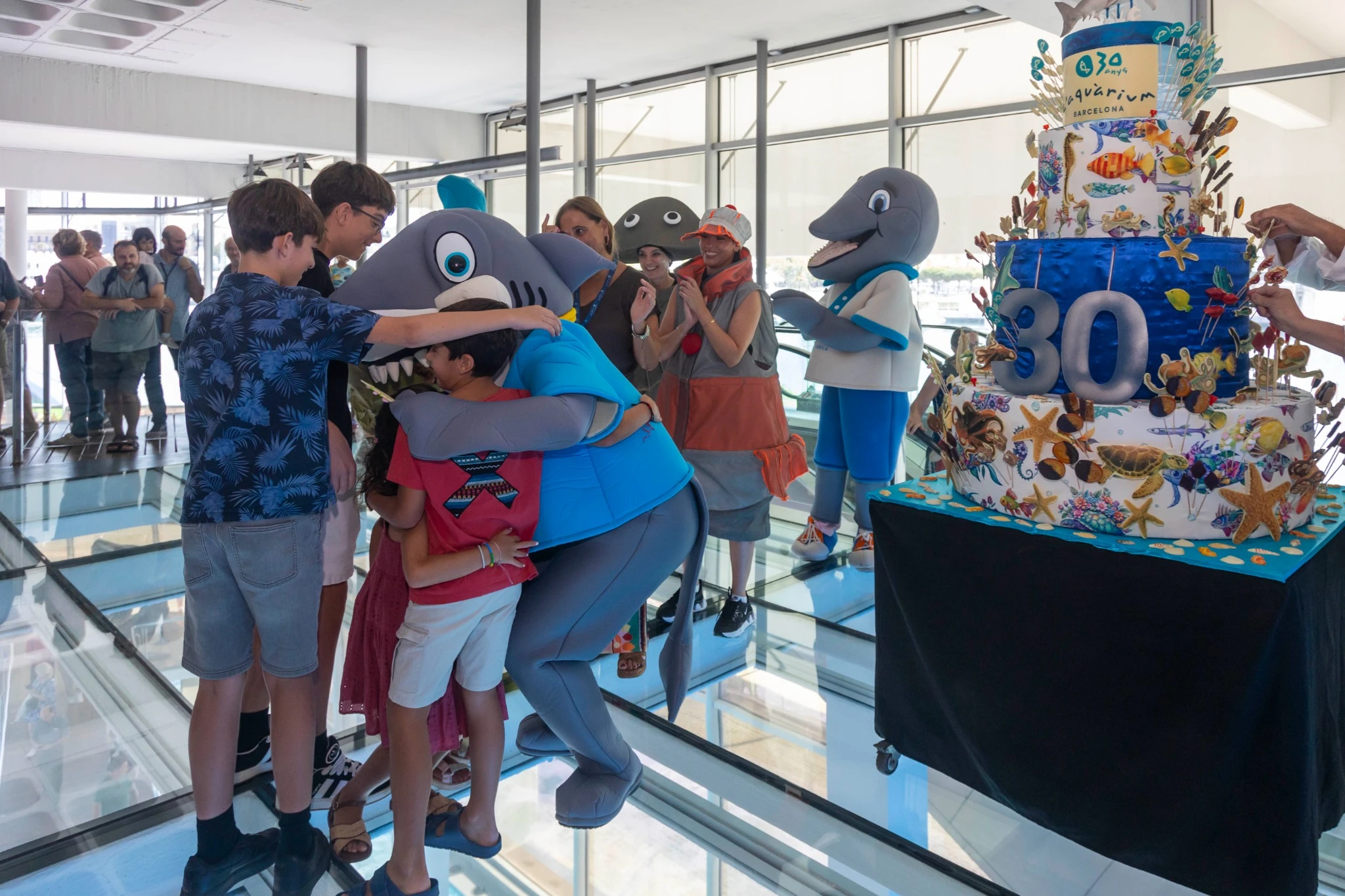 Niños y adultos celebran un cumpleaños junto a personajes marinos y una tarta temática en una pasarela de L’Aquàrium.